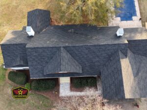 Bravo Company Roofing Aerial view of a house with a dark shingle roof, two chimneys, front porch, and landscaped yard; logo in lower left corner; pool visible in top right.