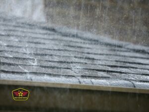 Bravo Company Roofing Heavy rain pours onto a shingled roof, creating visible splashes and wet surfaces. An emblem or badge is visible in the lower left corner of the image.
