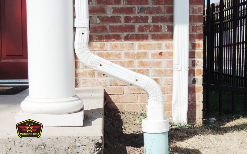 Bravo Company Roofing A white downspout attached to a brick wall drains into a green pipe in the ground next to a porch column. A logo in the lower left corner reads "Great Country Network.