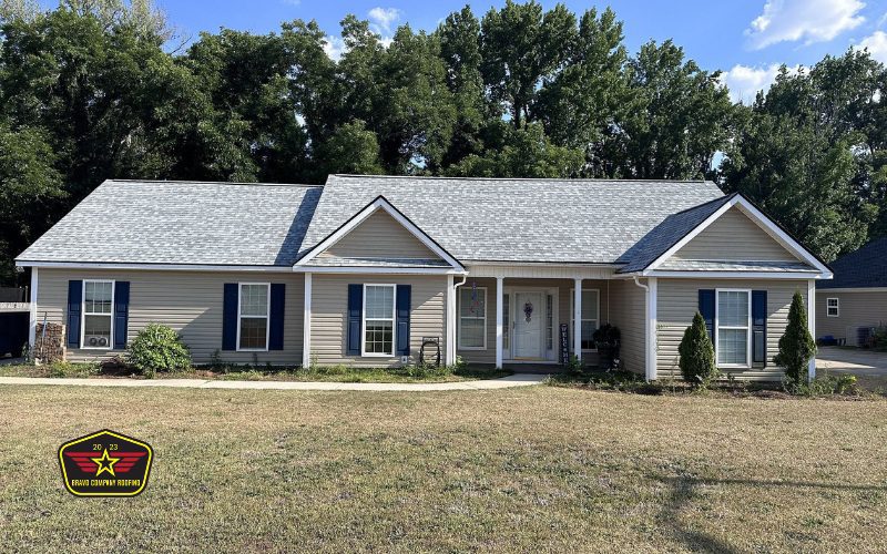 Bravo Company Roofing Single-story beige house with a gray roof, blue shutters, front porch, and a patchy lawn. Trees and a blue sky are in the background. A "Great Country Homes" logo is in the bottom left corner.