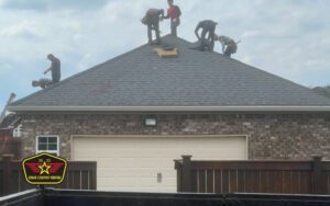 Bravo Company Roofing Five workers are installing or repairing shingles on the roof of a brick house. A company logo is visible in the lower left corner of the image.