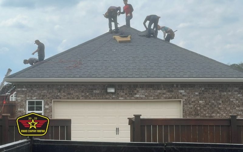 Bravo Company Roofing Five workers are installing or repairing shingles on the roof of a brick house. A company logo is visible in the lower left corner of the image.