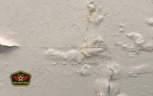 Bravo Company Roofing A white wall with cracked and peeling paint, surface bubbling, and visible water damage; a company logo is in the lower left corner.