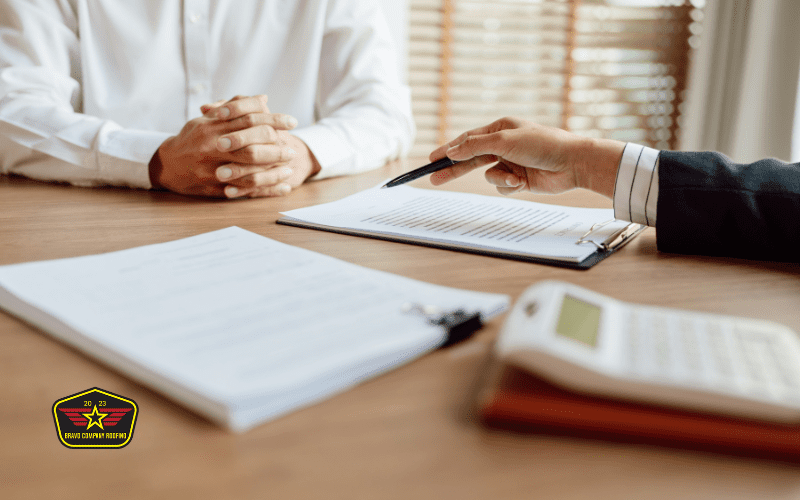 Bravo Company Roofing Two people sit at a desk with documents, a pen, and a calculator. One person gestures with a pen, while the other sits with hands clasped. A badge with a star logo is visible.