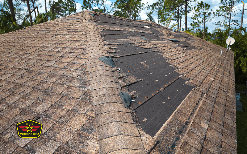 Bravo Company Roofing A house roof with missing and damaged shingles exposing the underlayment; trees and sky are visible in the background. A logo is present in the lower left corner.